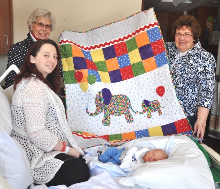 Gloversville Sew Busy Quilt Guild quilters Joanne Gasner, left, and Nancy Frank hold up a quilt they presented to Littauer newborn Asher Killian Betters, together with his mother Jackie Betters. The hand-crafted quilt was specially made for National Quilting Day